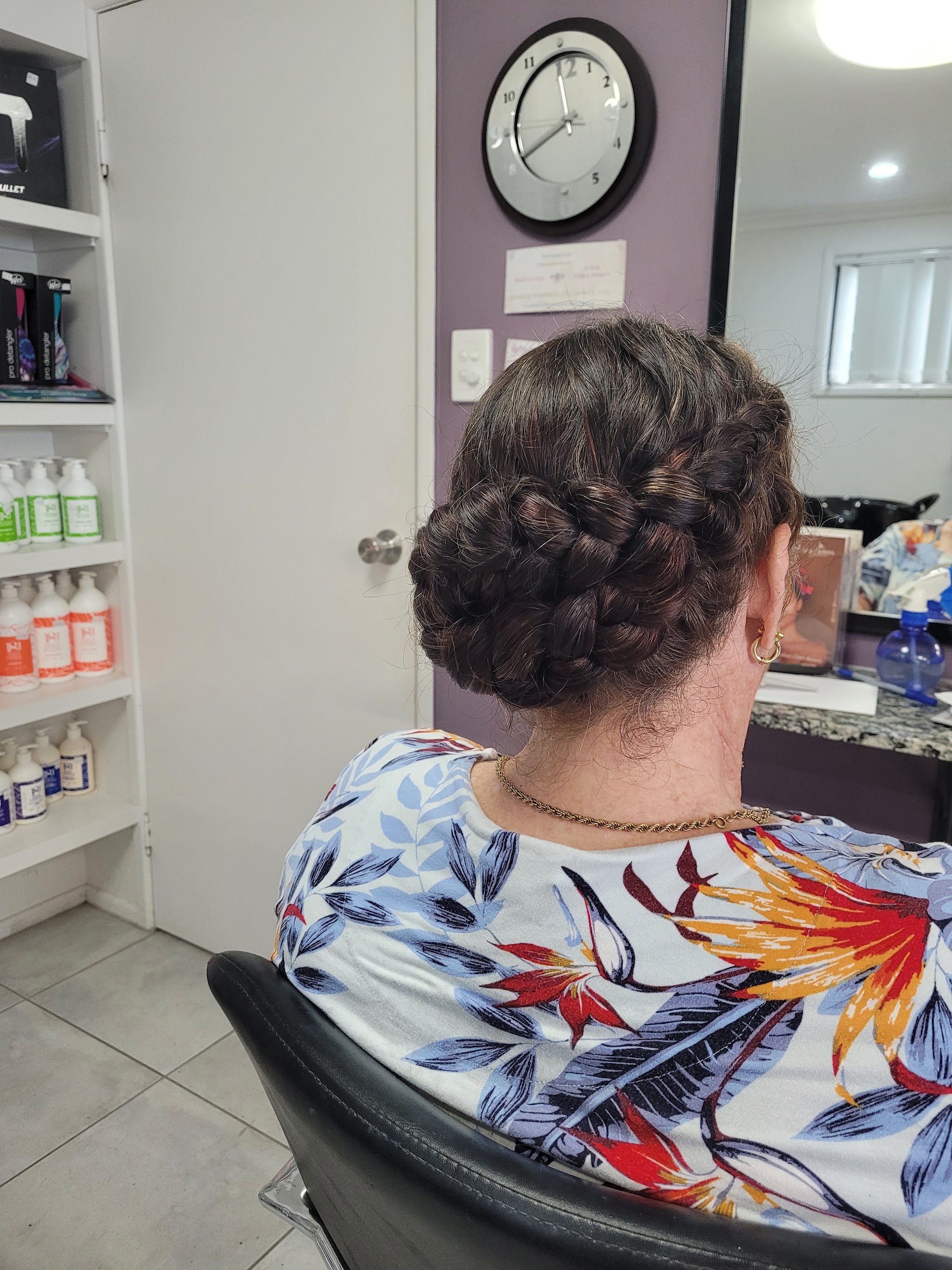 Inside the salon: a client in the chair showing off a braided low bun, with product shelves and the purple feature wall behind